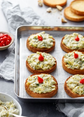 Cheesy Artichoke Crostini toasts arranged on a silver baking sheet with a bowl of artichoke topping and pepper drops on the side.