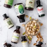 Nine bottles of different brands of pistachio extract, flat lay on a marble background with a bowl of pistachios.