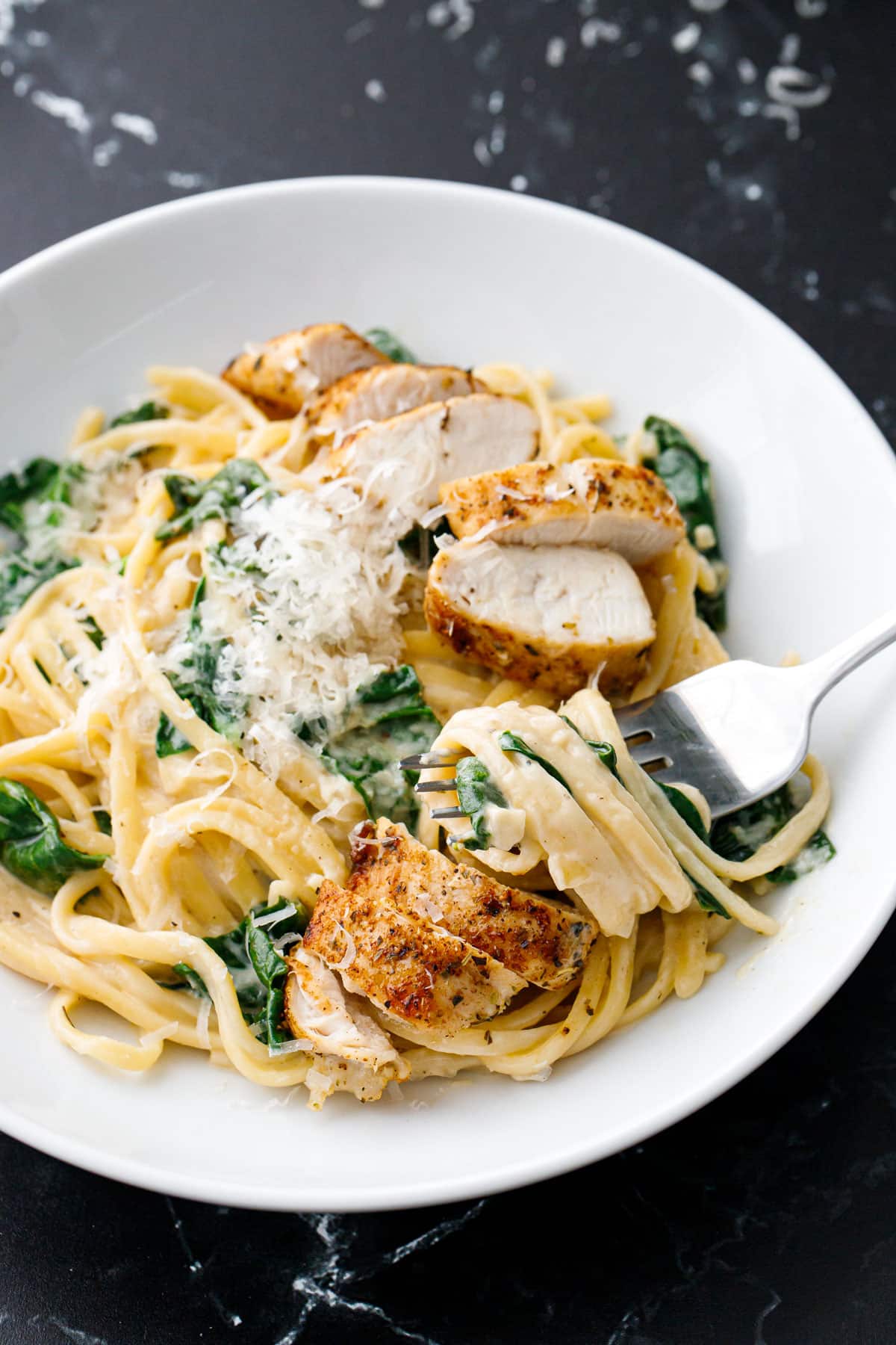 Fork with a twirl of linguine pasta showing the creamy sauce, plus visible bits of wilted spinach and pieces of seasoned chicken mixed in.