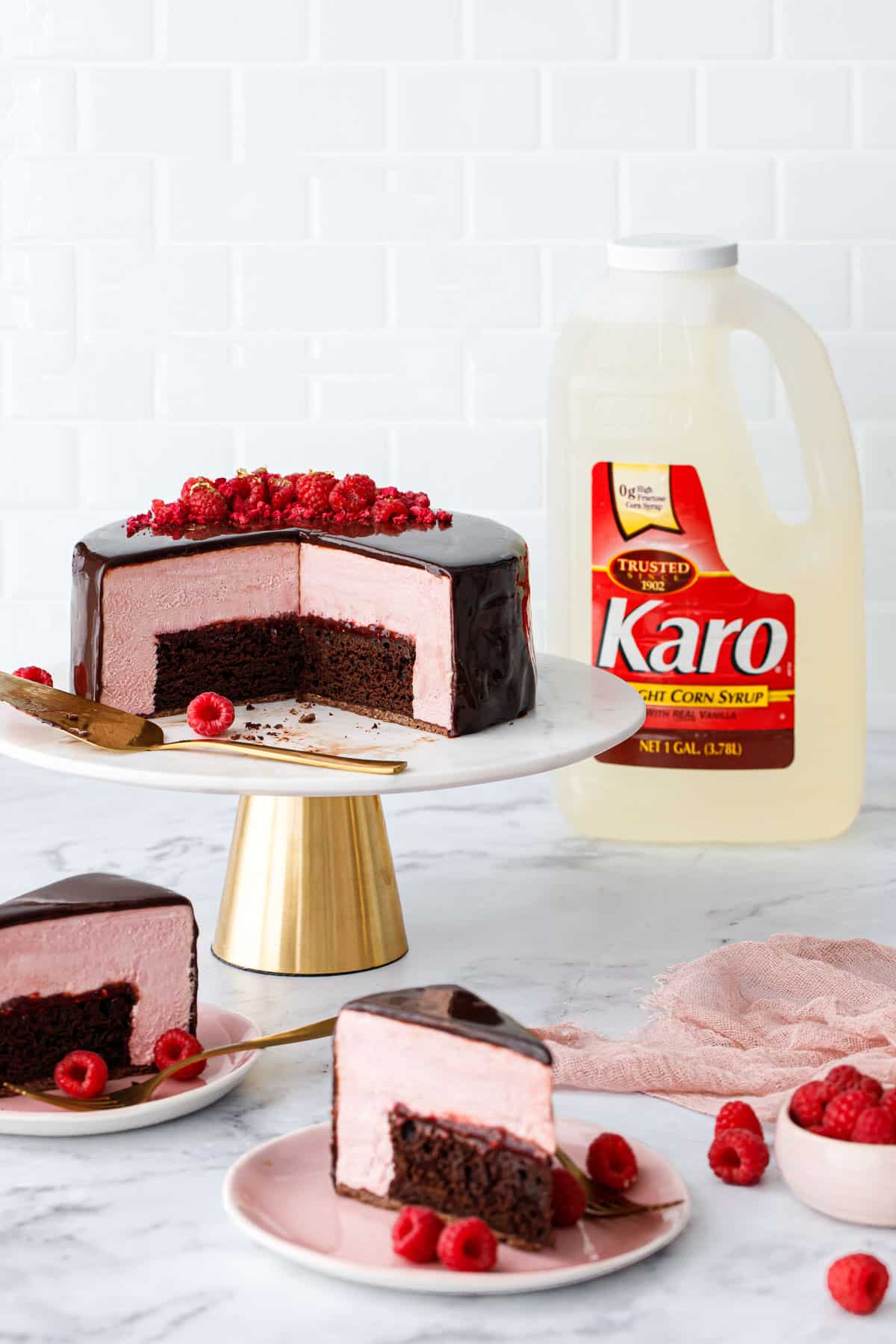 Slices of Chocolate Raspberry Mousse Cake on pink plates and full cake on a marble and gold cake stand, 1 gallon bottle of Karo corn syrup in the background.