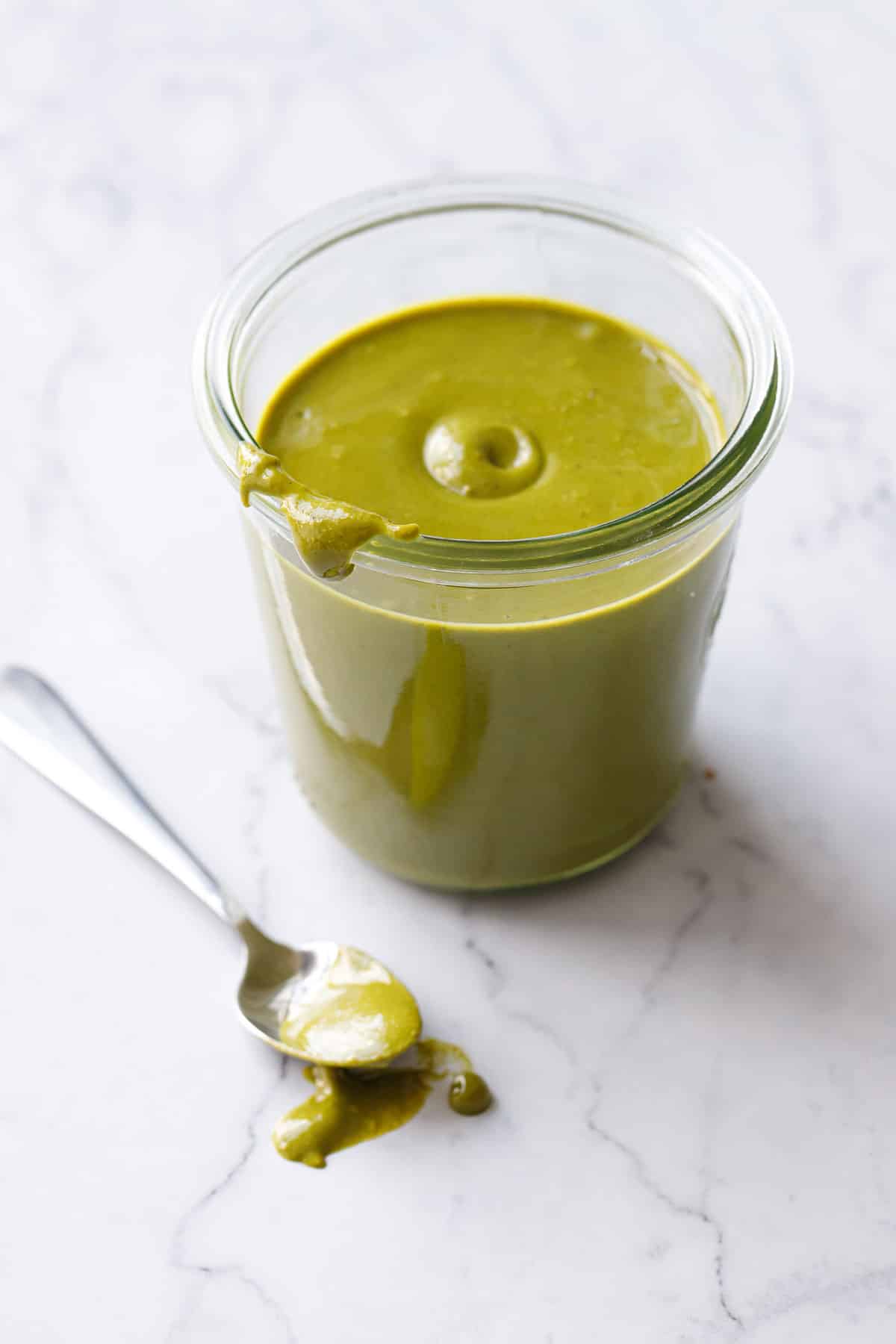 Glass jar of bright green Homemade Pistachio Butter on a marble background, spoonful oozing on the side showing the creamy texture.
