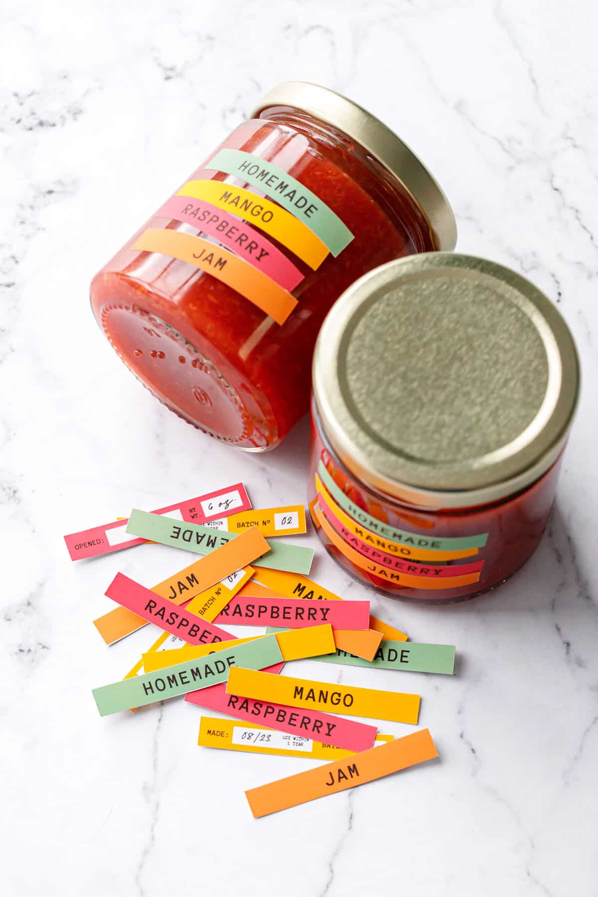 Sealed jars of Mango Raspberry Jam with custom colorful designed printed labels, some labels scattered on the background before being applied to jars.