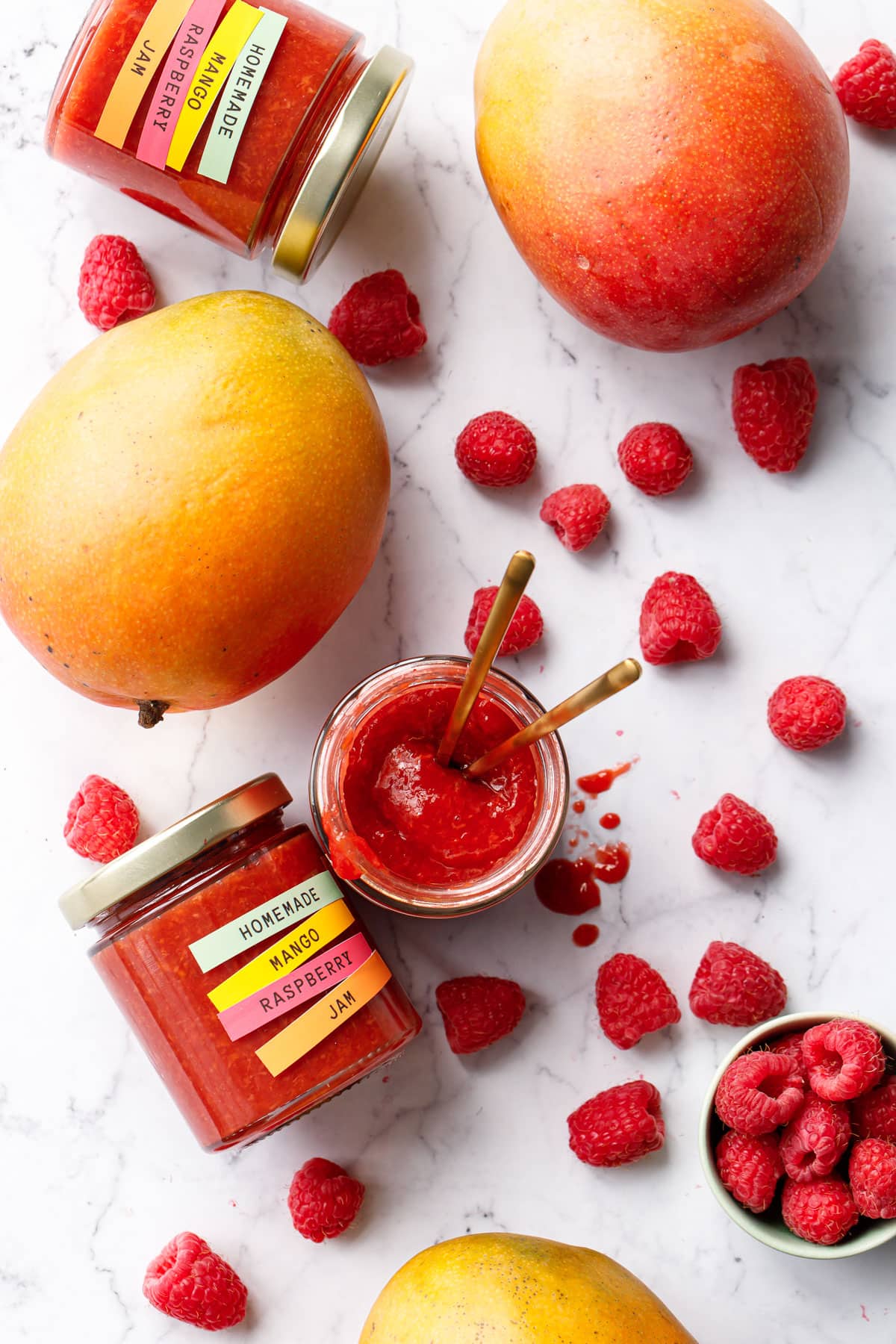 Overhead on a marble background, jars of Mango Raspberry Jam and fresh mangoes and raspberries scattered around.