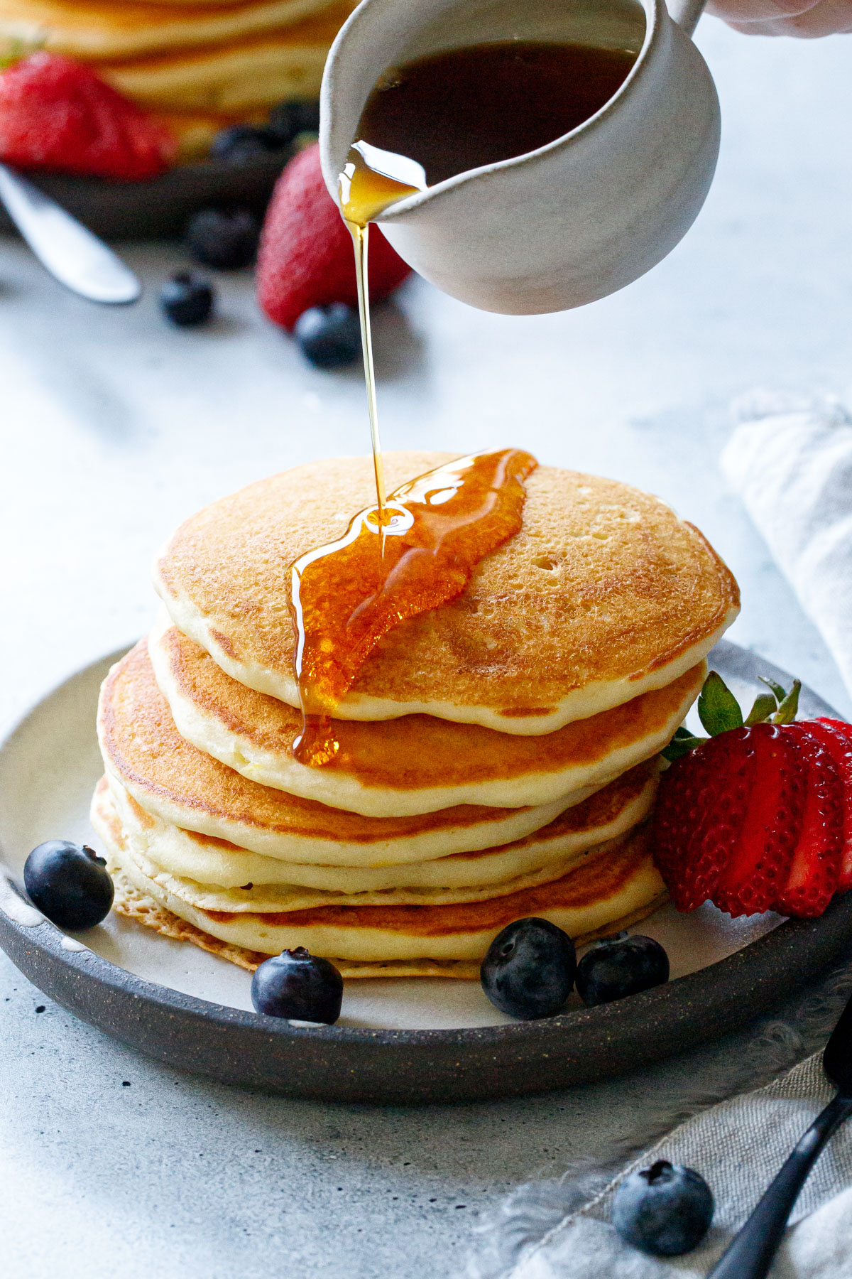 Pouring maple syrup on a stack of Olive Oil Pancakes with a drip down the side, a few blueberries and a sliced strawberry on the side.