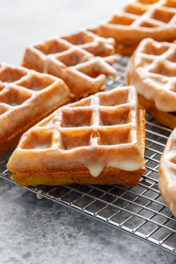 Closeup, one quarter round Pumpkin Donut Waffles with drips of Vanilla Cardamom Glaze down the side on a wire rack, gray background.