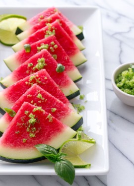 White plate with slices of watermelon, sprinkled with basil lime sugar. Garnished with fresh lime slices and a small bowl of sugar on the side.